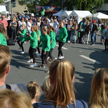 Tanzgruppe in gr&uuml;nen Jacken f&uuml;hrt Auff&uuml;hrung vor einer Menschenmenge auf einer Stra&szlig;e bei einem Fest vor.