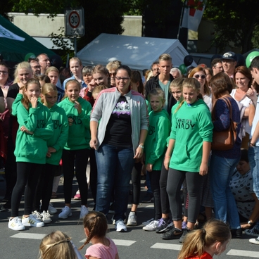Gruppierung von Menschen in gr&uuml;nen Sweatshirts bei einer Veranstaltung, umgeben von Zuschauern und St&auml;nden im Hintergrund.