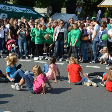 Gruppen von Menschen schauen einer Tanzauff&uuml;hrung auf einer Stra&szlig;e zu, w&auml;hrend Kinder in bunten T-Shirts sitzen.