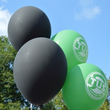 Drei Luftballons, zwei gr&uuml;n mit &sbquo;TUS&lsquo;-Logo und ein schwarzer, vor einem blauen Himmel und B&auml;umen im Hintergrund.