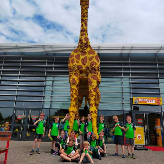 Gruppe von Kindern in gr&uuml;nen T-Shirts posiert vor einer gro&szlig;en Giraffe aus Legosteinen vor einem Geb&auml;ude.