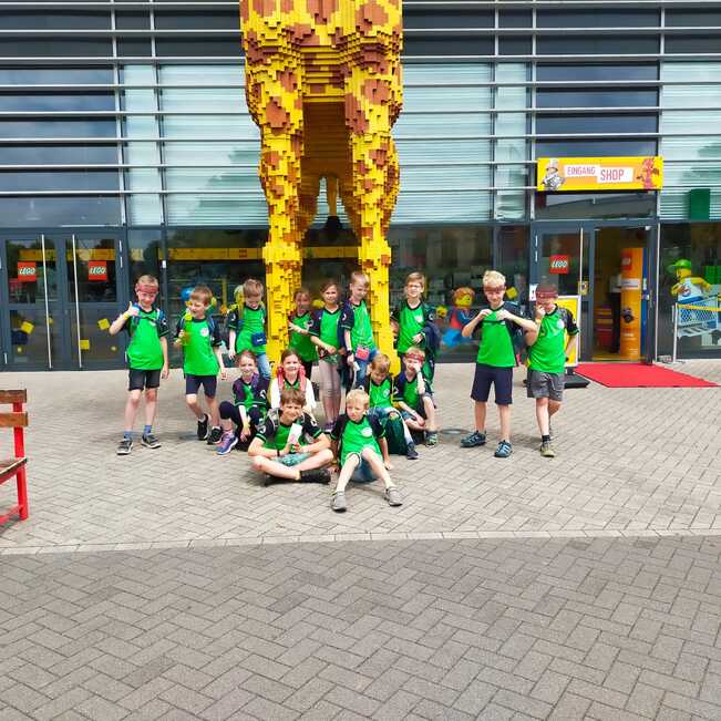 Gruppe von Kindern in gr&uuml;nen T-Shirts posiert vor einem LEGO-Geb&auml;ude mit gro&szlig;er LEGO-Skulptur.
