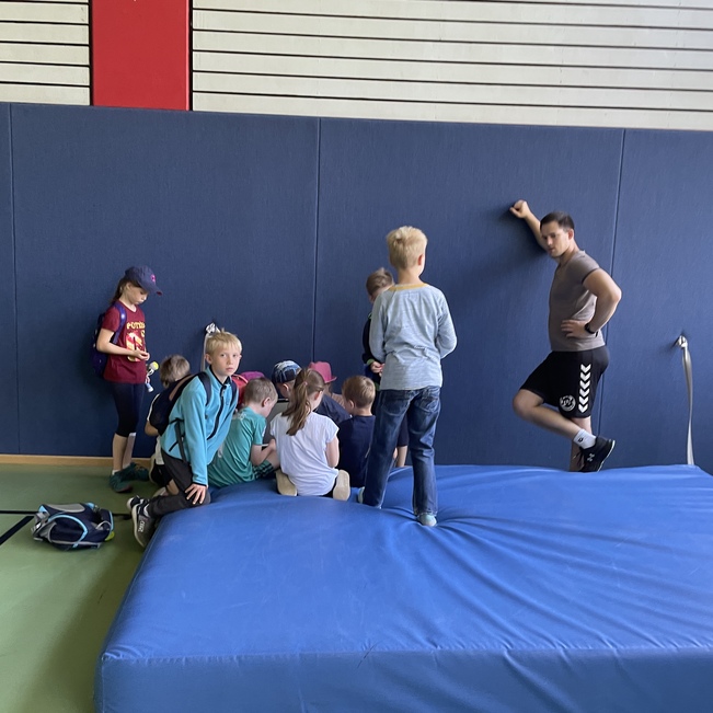 Gruppe von Kindern um einen blauen Turntisch versammelt, ein Junge lehnt an der Wand, Sportger&auml;te im Hintergrund sichtbar.
