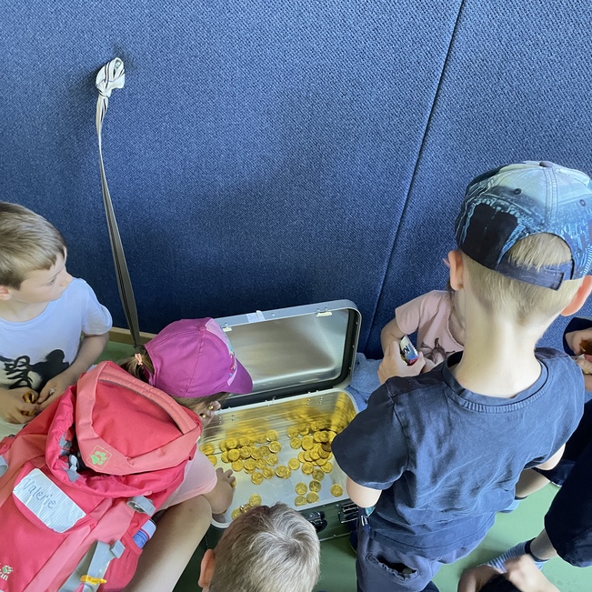 Gruppe von Kindern versammelt um eine Metallkiste mit Goldm&uuml;nzen auf einem gr&uuml;nen Boden.