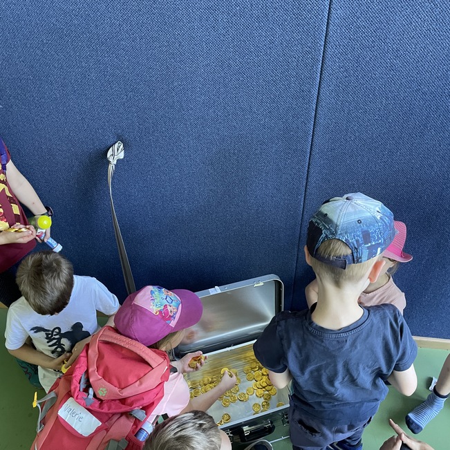 Gruppe von Kindern steht um einen offenen Koffer, der mit Spielzeugen gef&uuml;llt ist, an einer blauen Wand.