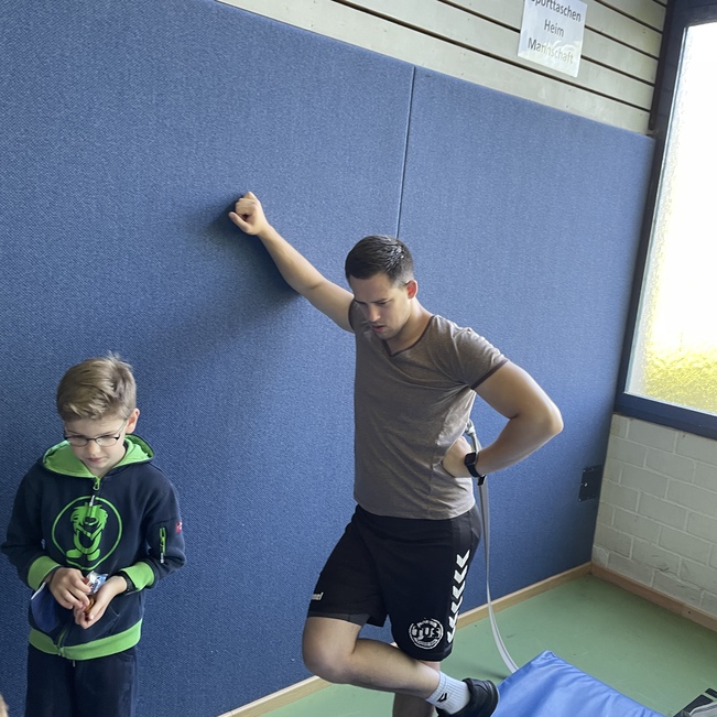 Ein Trainer steht an einer Wand und beobachtet einen Jungen, der mit einem Ball auf einer Turnmatte spielt.