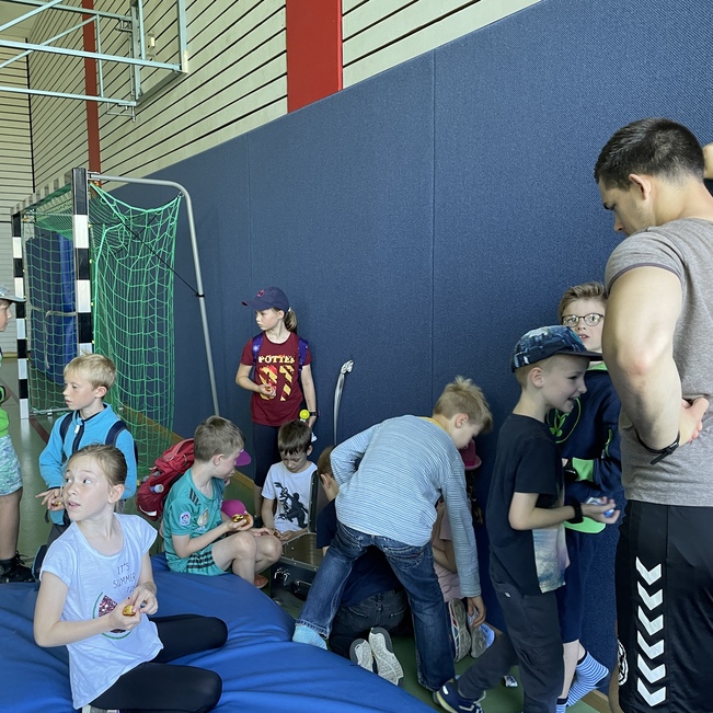 Gruppe von Kindern in einer Sporthalle, einige stehen, andere sitzen, ein Sportler spricht mit ihnen.