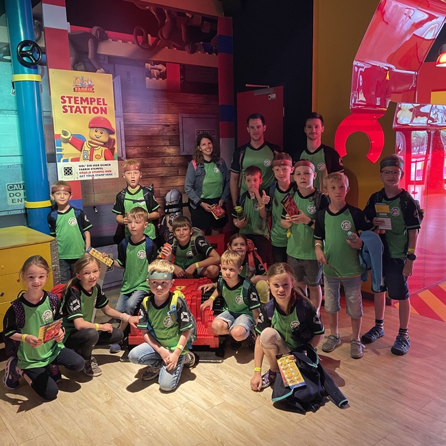 Gruppe von Kindern und Erwachsenen in Freizeitkleidung in einem bunten Indoor-Spielbereich vor einer Stempelstation.