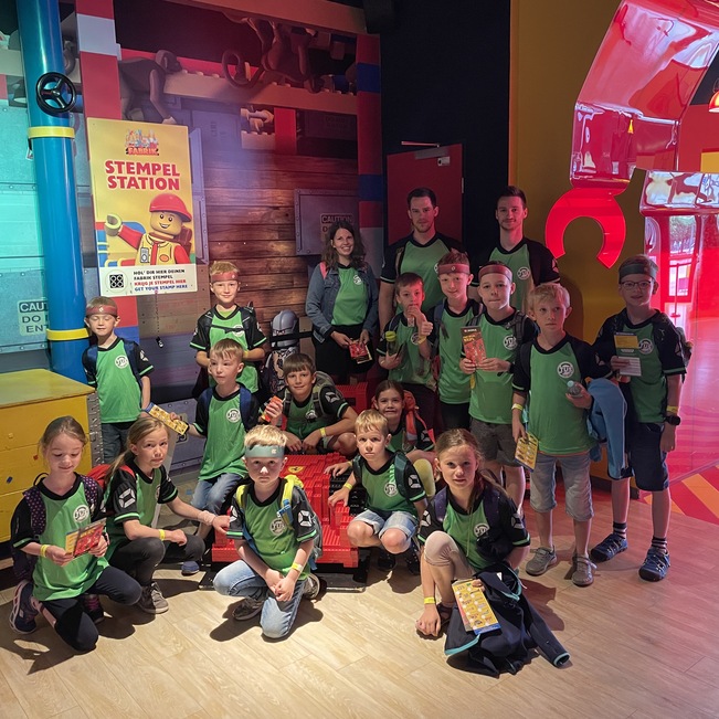 Gruppe von Kindern in gr&uuml;nen T-Shirts posiert vor einer farbenfrohen Spielumgebung mit Stempelstation.