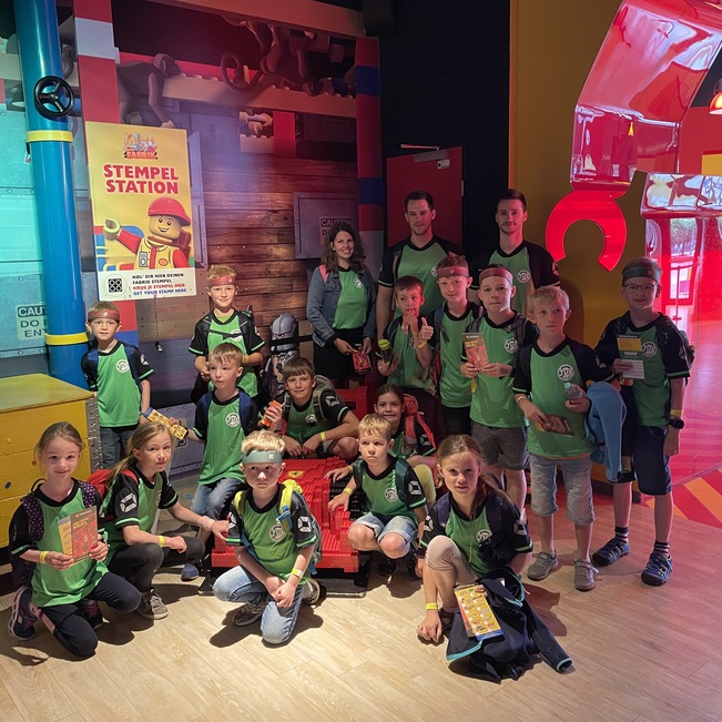 Gruppe von Kindern in gr&uuml;nen T-Shirts steht in einem bunten Raum mit Spielstationen und h&auml;lt Stempelkarten in der Hand.