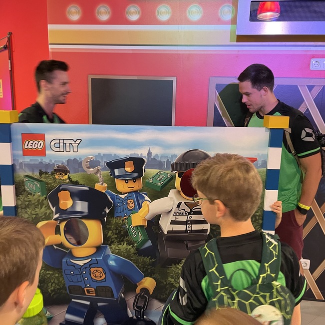 Zwei Erwachsene halten ein LEGO City-Plakat, umgeben von Kindern, die interessiert zuschauen.