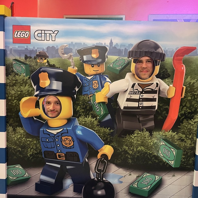 Zwei Personen posieren hinter einer LEGO-City Kulisse mit einer Polizei- und Verbrecherfigur, umgeben von Geldscheinen.