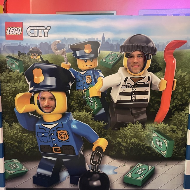 Zwei Personen posieren hinter einem LEGO City Fotohintergrund mit Polizisten und einem Gefangenen zwischen Geldscheinen.