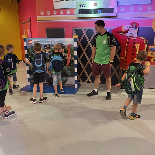 Gruppe von Kindern mit Rucks&auml;cken steht vor einer bunten Wandinstallation in einem Freizeitpark. Ein Betreuer erkl&auml;rt.