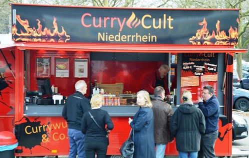 Lebensmittelausgabewagen mit der Aufschrift &bdquo;CurryCult Niederrhein&ldquo; und mehreren Warteg&auml;sten davor.