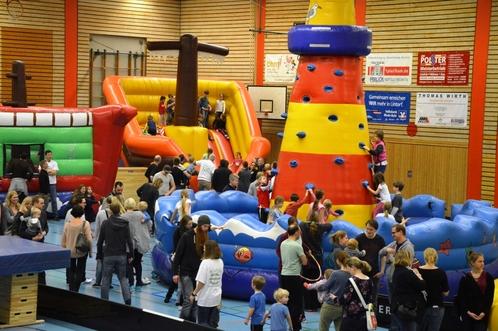Bunte H&uuml;pfburgen und Kletterw&auml;nde in einer vollen Sporthalle mit Kindern und Familien beim Spielen.