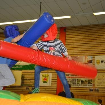 Zwei Kinder k&auml;mpfen auf einer aufblasbaren Kampfplattform mit Schaumstoffst&auml;ben in rot und blau.