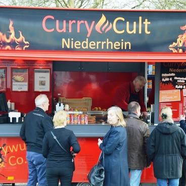 Lebensmittelausgabewagen mit der Aufschrift &bdquo;CurryCult Niederrhein&ldquo; und mehreren Warteg&auml;sten davor.