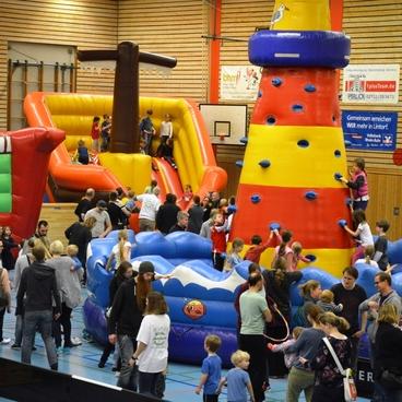 Bunte H&uuml;pfburgen und Kletterw&auml;nde in einer vollen Sporthalle mit Kindern und Familien beim Spielen.