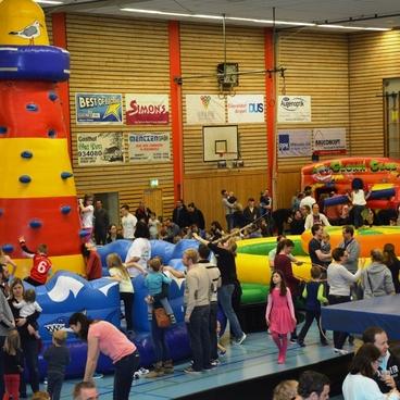 Buntes Indoor-Spielareal mit H&uuml;pfburgen und Spielenden, viele Menschen in einer sportlichen Halle.