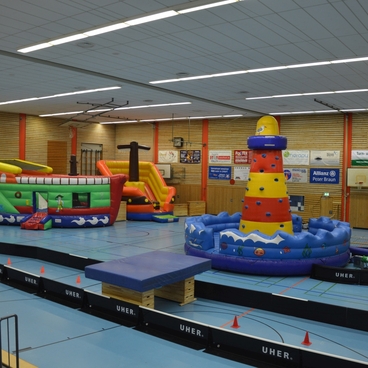 Bunte aufblasbare Spielger&auml;te in einer Sporthalle, darunter eine Rutsche und ein Wasser-Spielplatz.