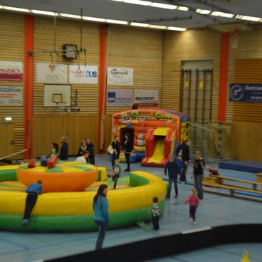 Buntes Trampolin und H&uuml;pfburg mit Kindern in einer Sporthalle, umgeben von Spielger&auml;ten und aktiven Besuchern.