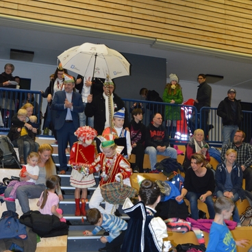 Karnevalsumzug mit verkleideten Kindern und Erwachsenen, die auf Stufen stehen und zuschauen. Ein Regenschirm wird gehalten.