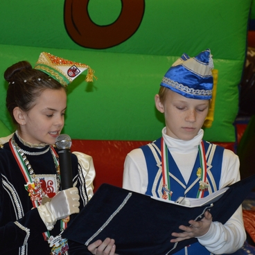 Zwei Kinder in Festkleidung stehen auf einer H&uuml;pfburg, eines spricht ins Mikrofon, das andere h&auml;lt ein Buch.