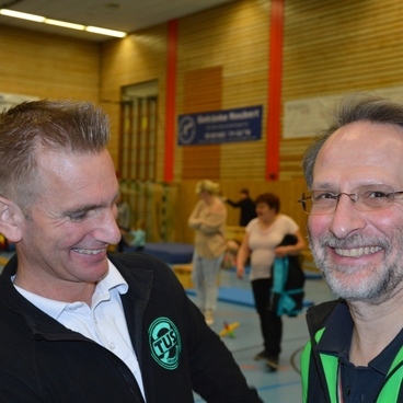 Zwei M&auml;nner l&auml;cheln und sprechen in einer Sporthalle, im Hintergrund sind weitere Personen und Turnger&auml;te zu sehen.