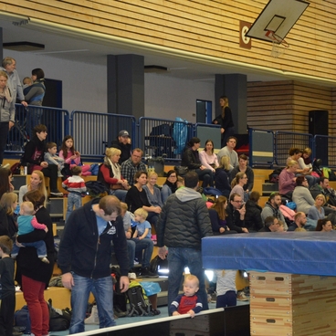 Zuschauer sitzen und stehen in einer Sporthalle, w&auml;hrend sie ein Turnereignis beobachten. Bunte Kleidung &uuml;berall.