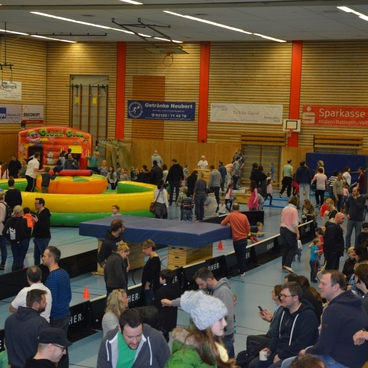 Bunte Veranstaltung in einer Sporthalle mit vielen Besuchern, H&uuml;pfburgen und aktiv spielenden Kindern auf einer Matte.