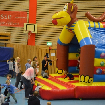 Kinder spielen auf einer gro&szlig;en H&uuml;pfburg in einer Sporthalle, begleitet von mehreren Erwachsenen.