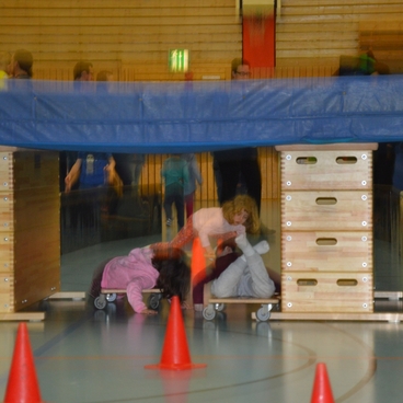 Kinder, die unter einer Turnbank durchrollen, umgeben von orangefarbenen H&uuml;tchen in einer Sporthalle.