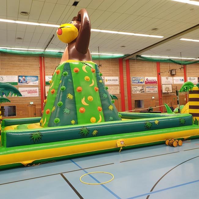 Bunte H&uuml;pfburg mit einem Affen auf einem H&uuml;gel, umgeben von palm&auml;hnlichen Pflanzen in einer Sporthalle.