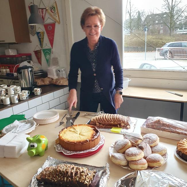 Frau mit kurzem Haar schneidet Kuchen auf einem Tisch mit mehreren Backwaren und Tassen im Hintergrund.