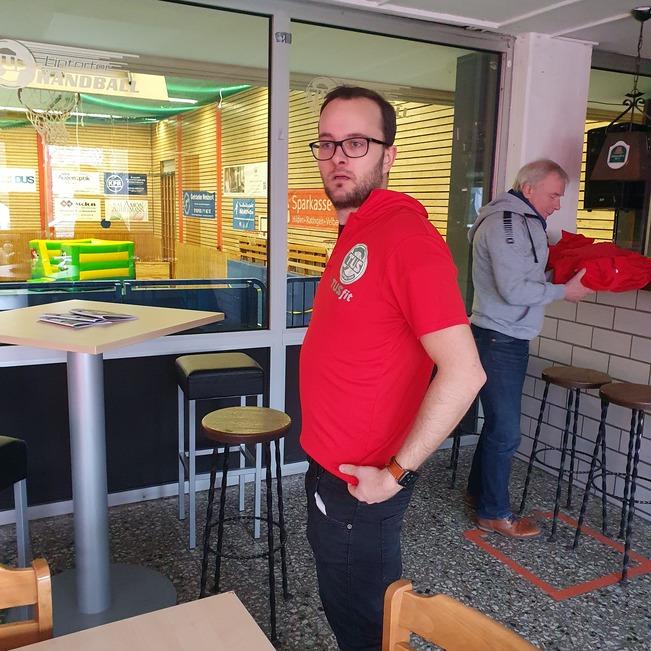 Mann in rotem Shirt steht an einem Bartisch in einer Sporteinrichtung, im Hintergrund ein Handballfeld.