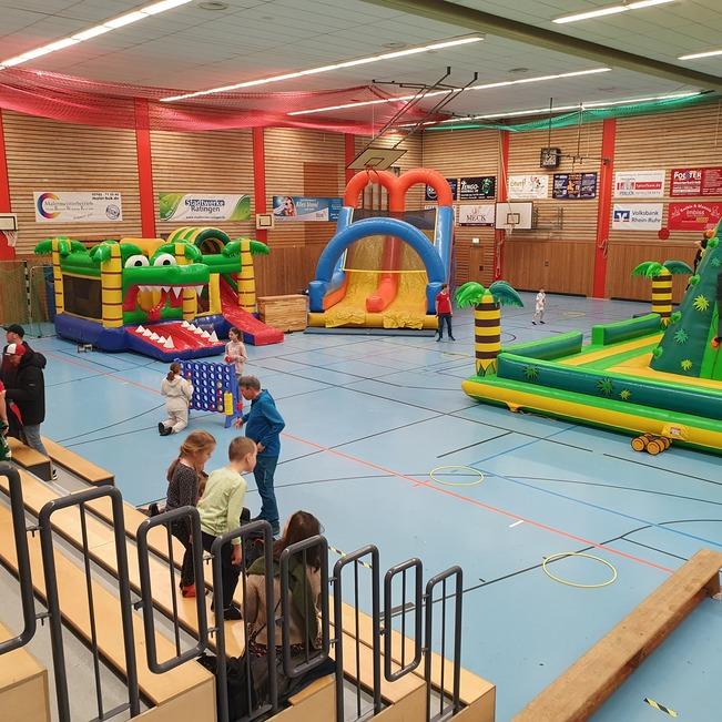 Blick in eine Sporthalle mit mehreren bunten H&uuml;pfburgen und Kindern, die darauf spielen und sich umsehen.