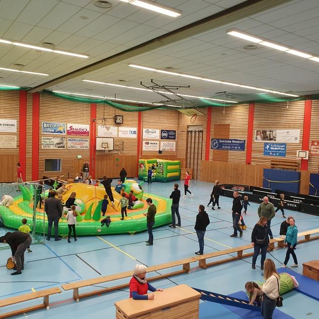 Kinder spielen in einer Sporthalle auf einem gro&szlig;en, gr&uuml;nen H&uuml;pfburgen-Parcours mit verschiedenen Spielstationen.
