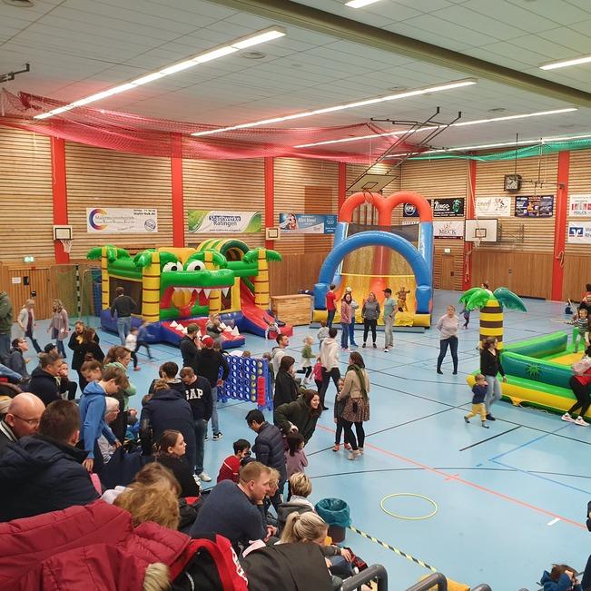 Belebte Sporthalle mit mehreren H&uuml;pfburgen und zahlreichen Kindern und Erwachsenen, die spielen und zuschauen.