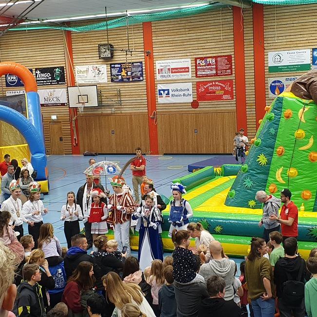 Bunt dekorierte B&uuml;hne mit Menschen in Kost&uuml;men und H&uuml;pfburg w&auml;hrend einer Veranstaltung in einer Sporthalle.