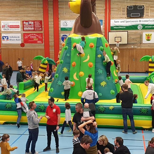 Kinder spielen auf einer gro&szlig;en, bunten H&uuml;pfburg mit Kletterwand und tropischer Palmenumgebung in einer Sporthalle.