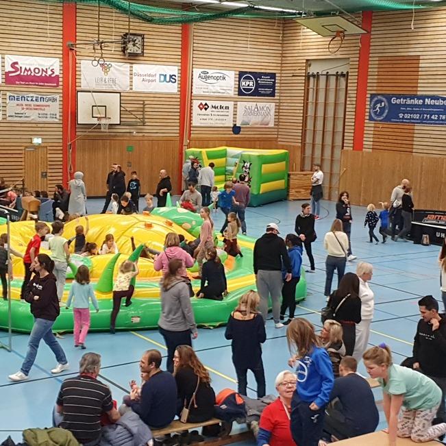 Belebte Turnhalle mit einer gro&szlig;en H&uuml;pfburg und vielen Kindern, die spielen, sowie Zuschauern auf B&auml;nken.