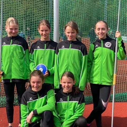 Sechs M&auml;dchen in gr&uuml;nen Trainingsjacken stehen und sitzen auf einer Sportanlage mit einem Wurfger&auml;t.