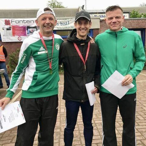 Drei M&auml;nner stehen l&auml;chelnd zusammen, jeder mit einem Zertifikat in der Hand, vor einer Veranstaltungsb&uuml;hne im Freien.