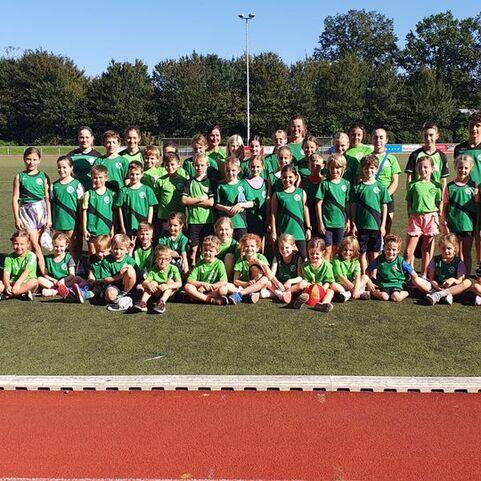 Gruppenspiel von Kindern und Jugendlichen in gr&uuml;nen Trikots auf einem Sportplatz, umgeben von B&auml;umen.