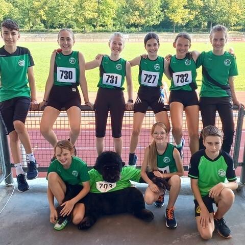 Gruppe von jungen Athleten in Sportkleidung mit Startnummern, posierend an einer Sportanlage mit gr&uuml;ner Rasenfl&auml;che.