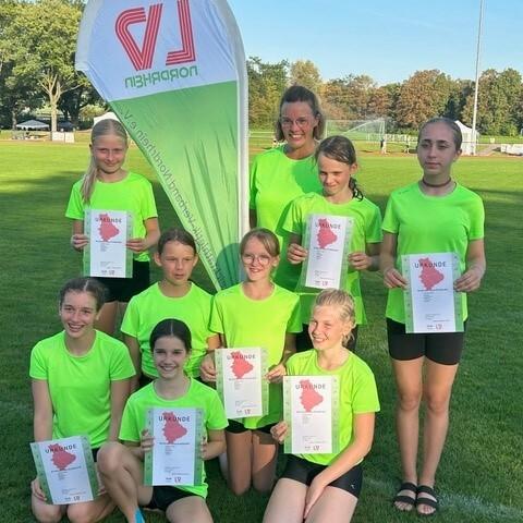 Gruppe von M&auml;dchen in neonfarbenen T-Shirts h&auml;lt Urkunden auf einem Sportplatz, im Hintergrund ein Vereinsbanner.