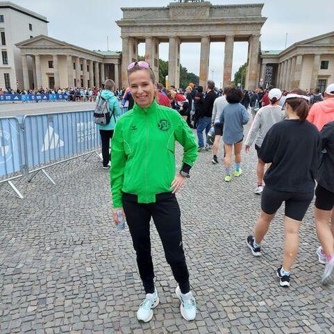 L&auml;chelnde Person in gr&uuml;ner Jacke steht vor dem Brandenburger Tor, umgeben von Laufbegeisterten bei einer Veranstaltung.