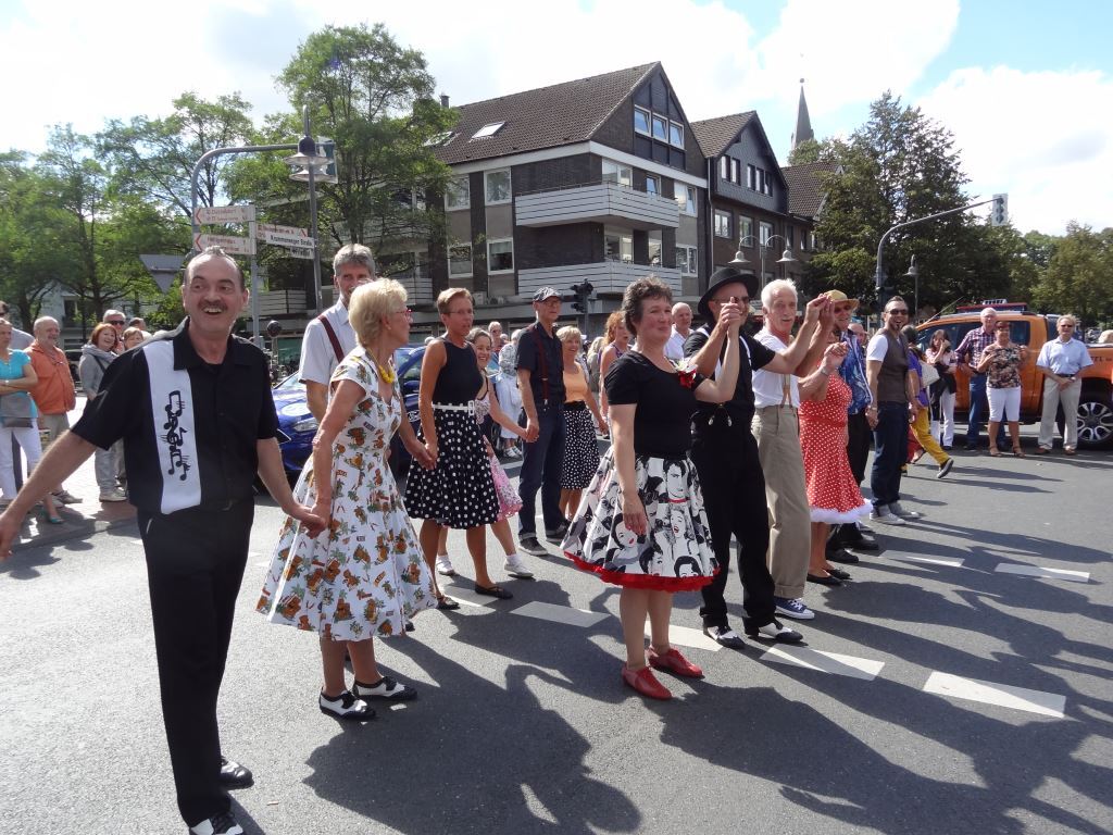Tanzgruppe in bunten Kleidern auf der Stra&szlig;e, umgeben von Zuschauern bei einem Festumzug.
