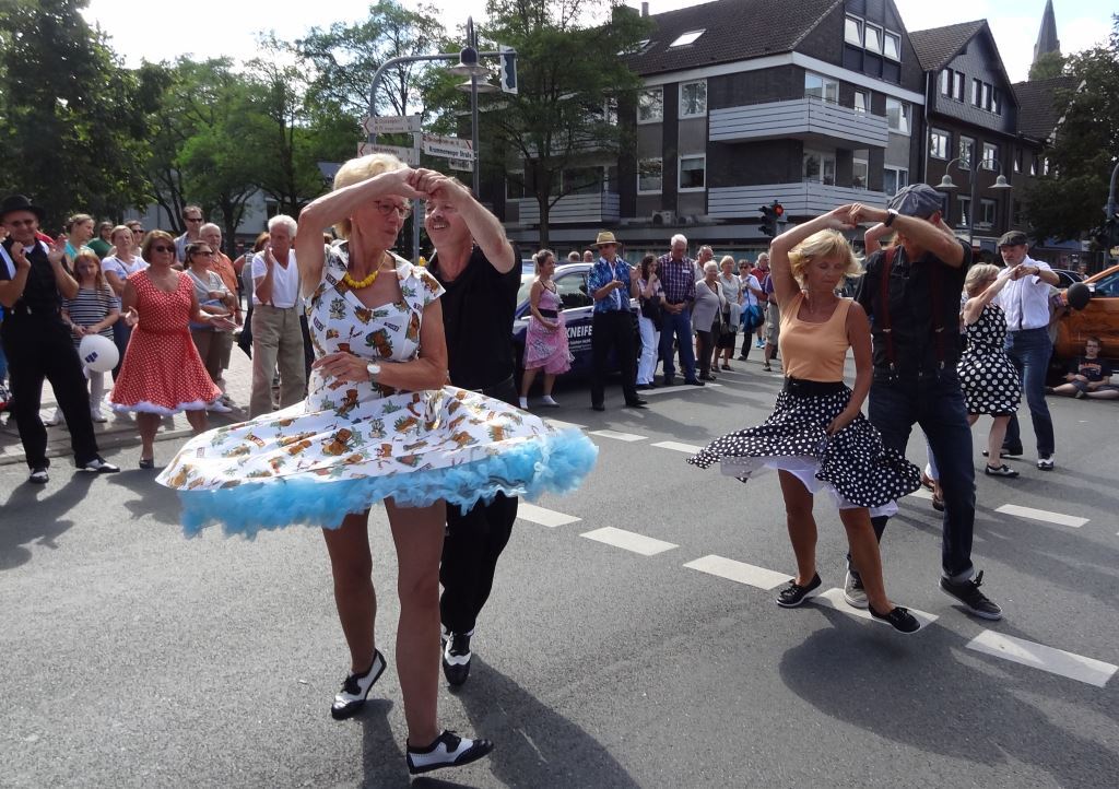 Tanzende Paare in bunten Kleidern f&uuml;hren einen Lindy Hop auf der Stra&szlig;e vor einer Menge Schaulustiger auf.